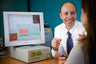 A man in a white shirt and blue tie sits at a desk smiling while talking to a woman, with a computer monitor showing dental imaging software and an oral chromo device on the desk in an office setting.
