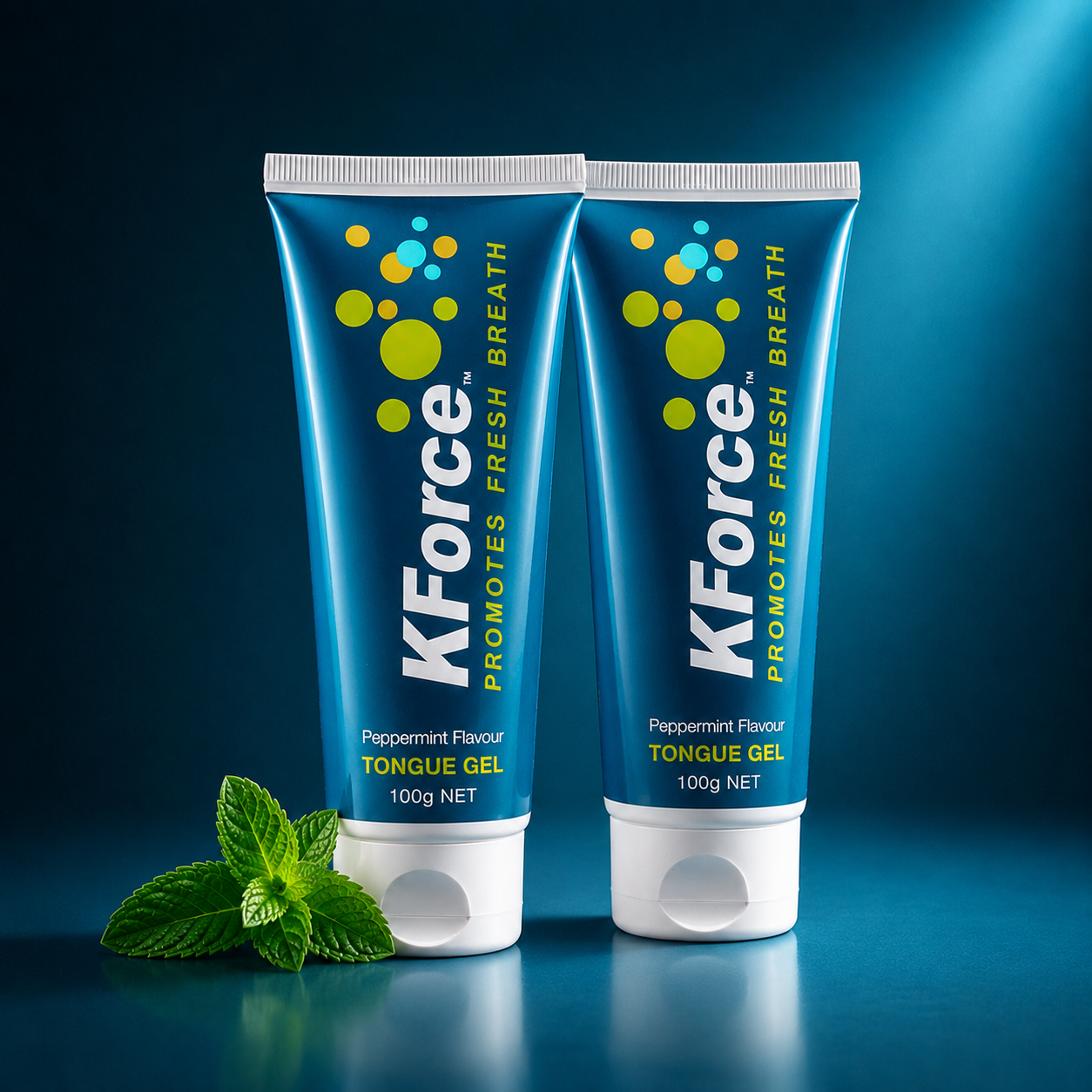 Two tubes of KForce Peppermint Flavour Tongue Gel standing upright on a dark blue reflective surface, with a small mint leaf placed in front. The tubes are blue with yellow and white graphics and text, and the background is a dark blue studio setting with light beams.